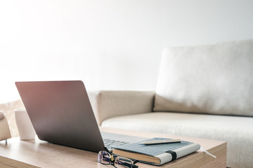Laptop computer, notebook and glasses on the wood table with blurred couch background in living room.