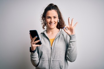 Fototapeta premium Beautiful sportswoman with curly hair listening to music using smartphone and earphones doing ok sign with fingers, excellent symbol