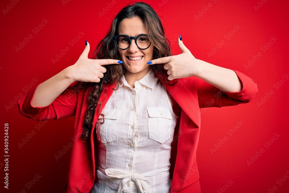 Sticker Young beautiful woman with curly hair wearing jacket and glasses over red background smiling cheerful showing and pointing with fingers teeth and mouth. Dental health concept.