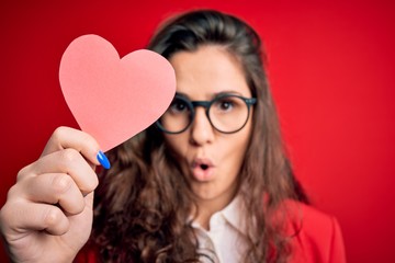 Young beautiful woman with curly hair holding paper heart over isolated red background scared in shock with a surprise face, afraid and excited with fear expression