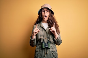 Young beautiful tourist woman on vacation wearing explorer hat and binoculars amazed and surprised...