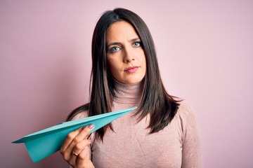 Young woman with blue eyes holding paper airplane standing over isolated pink background with a confident expression on smart face thinking serious