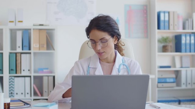 Medium shot of Caucasian female medical worker wearing medical gown and stethoscope sitting at desktop at laptop and prescribing pills to online patient