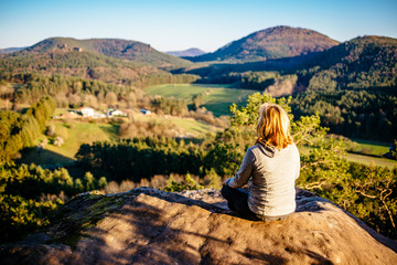 A blonde woman sits on a sandstone cliff in the Palatinate Forest and looks at the sunset