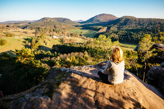 A blonde woman sits on a sandstone cliff in the Palatinate Forest and looks at the sunset