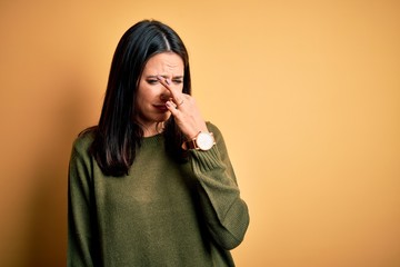 Young brunette woman with blue eyes wearing green casual sweater over yellow background smelling something stinky and disgusting, intolerable smell, holding breath with fingers on nose. Bad smell