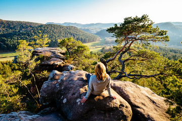 A blonde woman sits on a sandstone cliff in the Palatinate Forest and looks at the sunset - obrazy, fototapety, plakaty