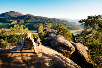 A blonde woman sits on a sandstone cliff in the Palatinate Forest and looks at the sunset