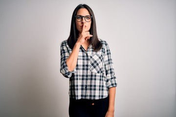 Young brunette woman with blue eyes wearing casual shirt and glasses over white background asking to be quiet with finger on lips. Silence and secret concept.