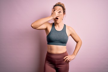Young beautiful blonde sportswoman doing sport wearing sportswear over pink background peeking in shock covering face and eyes with hand, looking through fingers with embarrassed expression.