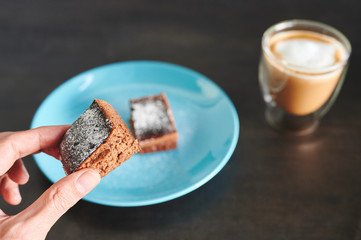 Detail of hand holding a piece of cake in the foreground with a coffee and more cake out of focus in the background