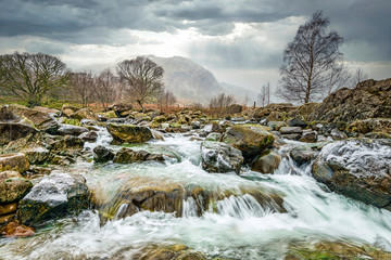 Stormy clouds and rain with a fluttering of fallen snow over Langstrath Beck in winter on the Cumbria Way footpath near Rosthwaite in Borrowdale, Lake District National Park, Cumbrian Mountains, Cumbr