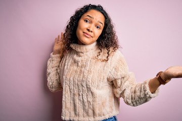 Young beautiful woman with curly hair wearing casual sweater standing over pink background clueless...