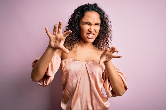 Young beautiful woman with curly hair wearing casual t-shirt standing over pink background smiling funny doing claw gesture as cat, aggressive and sexy expression