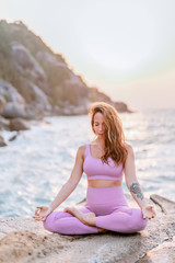 Fototapeta na wymiar Pregnant woman practices yoga, sitting in lotus position on the beach, on the rocks. Hands of girl in a pose meditating. Serenity practicing at sunrise, meditation. Woman in a pink-purple tracksuit.