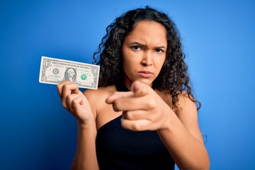 Young beautiful woman with curly hair holding one dollar over isolated blue background pointing with finger to the camera and to you, hand sign, positive and confident gesture from the front