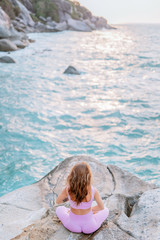 Pregnant woman practices yoga, sitting in lotus position on the beach, on the rocks. Hands of girl in a pose meditating. Serenity practicing at sunrise, meditation. Woman in a pink-purple tracksuit.