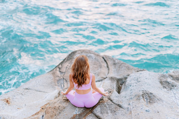Pregnant woman practices yoga, sitting in lotus position on the beach, on the rocks. Hands of girl in a pose meditating. Serenity practicing at sunrise, meditation. Woman in a pink-purple tracksuit.