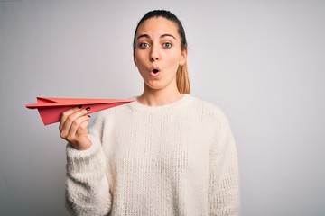 Beautiful blonde woman with blue eyes holding red paper airplane over white background scared in...