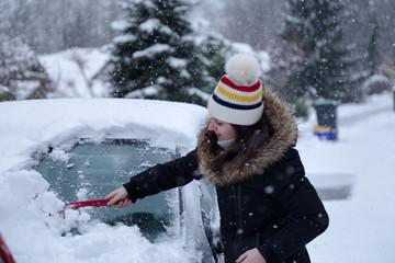 Young attractive brunette woman clearing snow off a car with a ice scraper in the winter. © Adam