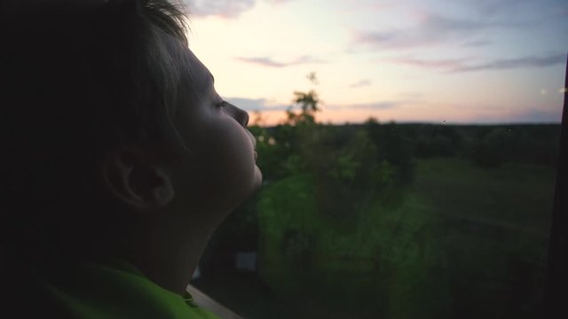 Closeup view profile video portrait of cute young kid standing near window and looking at beautiful evening sunset nature landscape seen behind glass.