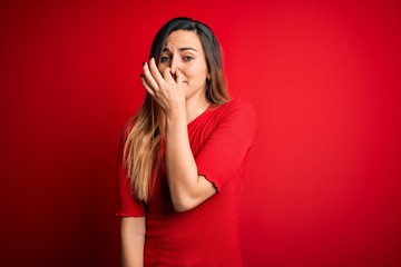 Young beautiful blonde woman with blue eyes wearing casual t-shirt over red background smelling something stinky and disgusting, intolerable smell, holding breath with fingers on nose. Bad smell