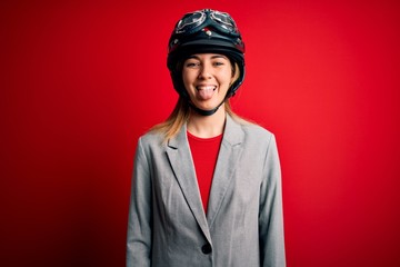 Young beautiful blonde motorcyclist woman wearing motorcycle helmet over red background sticking tongue out happy with funny expression. Emotion concept.