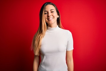 Beautiful blonde woman with blue eyes wearing casual white t-shirt over red background with a happy and cool smile on face. Lucky person.