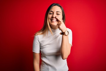 Beautiful blonde woman with blue eyes wearing casual white t-shirt over red background Pointing with hand finger to face and nose, smiling cheerful. Beauty concept