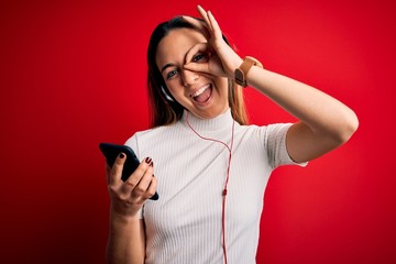Young beautiful brunette woman listening to music using smartphone and headphones with happy face smiling doing ok sign with hand on eye looking through fingers