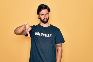 Young handsome hispanic volunteer man wearing volunteering t-shirt as social care looking unhappy and angry showing rejection and negative with thumbs down gesture. Bad expression.
