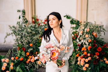original bride in a white trouser suit posing against the background of an unusual composition of fresh flowers in coral color for an outdoor wedding ceremony