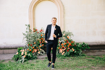 groom in a suit posing against the backdrop of an unusual composition of fresh flowers of coral color for an outdoor wedding ceremony