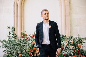 groom in a suit posing against the backdrop of an unusual composition of fresh flowers of coral color for an outdoor wedding ceremony