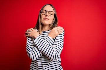 Young beautiful blonde woman with blue eyes wearing glasses standing over red background Hugging...