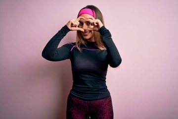 Young blonde fitness woman wearing sport workout clothes over isolated background Doing heart shape with hand and fingers smiling looking through sign