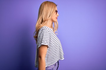 Young beautiful blonde woman wearing striped t-shirt and glasses over purple background looking to side, relax profile pose with natural face and confident smile.