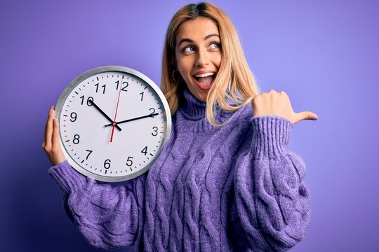 Young beautiful blonde woman doing countdown holding big clock over purple background pointing and showing with thumb up to the side with happy face smiling
