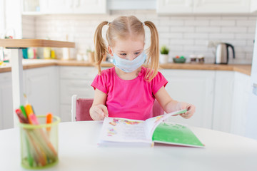 Little blonde girl with two ponytales reading a book at home in the kitchen wearing medical...