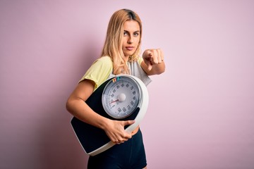 Young beautiful blonde sporty woman on diet holding weight machine over pink background pointing with finger to the camera and to you, hand sign, positive and confident gesture from the front