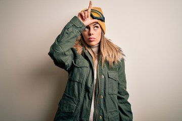 Young brunette skier woman wearing snow clothes and ski goggles over white background making fun of people with fingers on forehead doing loser gesture mocking and insulting.