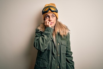 Young brunette skier woman wearing snow clothes and ski goggles over white background looking stressed and nervous with hands on mouth biting nails. Anxiety problem.