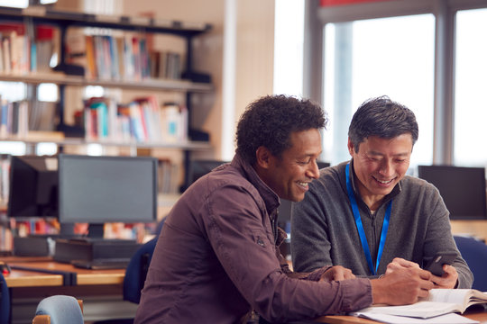Teacher With Mature Male Adult Student Using Mobile Phone At Table Working In College Library