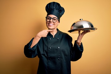 Young beautiful brunette chefwoman wearing cooker uniform and hat holding tray with dome very happy...