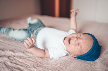Obraz na płótnie Canvas Cute crying little baby boy lying on pink blanket at bedroom. Newborn. Baby stays awake on the bed. Closeup portrait of emotional newborn baby in blue jeans, blue cap, and white t-shirt