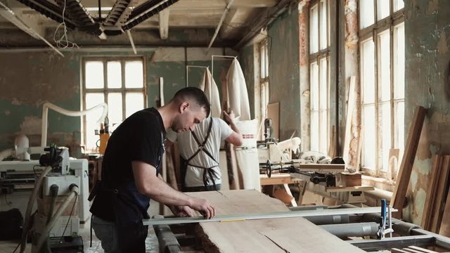 Two men working in wood factory. Professional carpenter makes wooden table in industrial interior. Woodworking and craftmanship concept