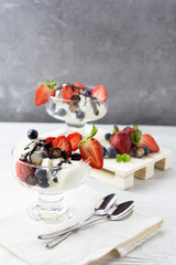 Two servings of vanilla ice cream in a glass bowl, decorated with strawberries and blueberries, drenched with dark chocolate. Served for a couple on a table with spoons and some berries. Vertical.