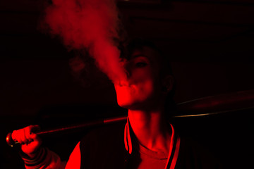 Contemporary female in bomber jacket holding a black baseball bat on shoulder while smoking with red light on background