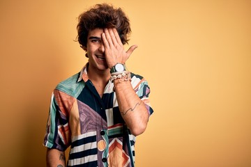 Young handsome man on vacation wearing summer shirt over isolated yellow background covering one eye with hand, confident smile on face and surprise emotion.