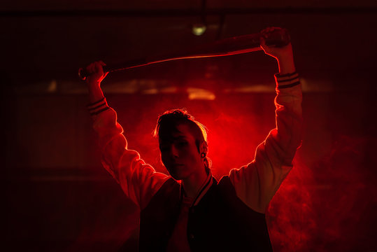Rebel woman in casual jacket with piercing and modern hairstyle holding bat among colorful red light and steam